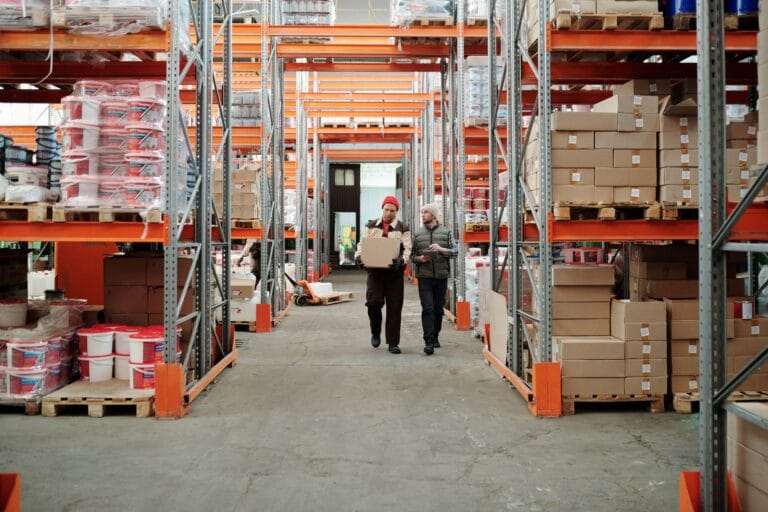 Two workers handle a package in a spacious warehouse surrounded by shelves stocked with boxes and products.